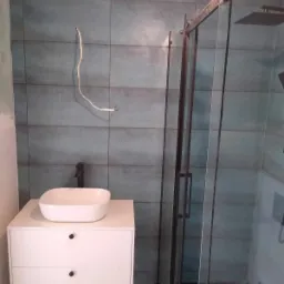 Niedokończona łazienka z niebiesko-szarymi płytkami, białą szafką z umywalką nablatową, kabiną prysznicową z czarną ramą i wystającym przewodem elektrycznym.