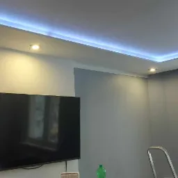 Szare wnętrze pokoju z podświetlanym sufitem LED, widoczny fragment telewizora, okno i drabina oparta o ścianę.