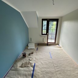 Usługi Budowlane Artur Ochodek - Pokój w trakcie malowania: jedna ściana pomalowana na niebiesko, podłoga zabezpieczona papierem malarskim, widoczne puszki z farbą i akcesoria malarskie. Na suficie widoczny hak.
