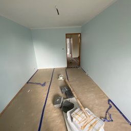 Usługi Budowlane Artur Ochodek - Pokój w trakcie malowania na jasny, błękitny odcień, podłoga zabezpieczona papierem malarskim, widoczne narzędzia i materiały malarskie, w tle otwarte drzwi do kolejnego pomieszczenia.