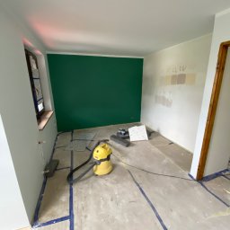 Usługi Budowlane Artur Ochodek - Pokój w trakcie prac: zielona ściana, próbki farb na sąsiedniej ścianie, zabezpieczona podłoga taśmą, odkurzacz przemysłowy i fragmenty płytek podłogowych.