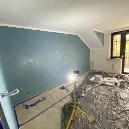 Usługi Budowlane Artur Ochodek - Pokój w trakcie malowania na niebiesko, zabezpieczony folią i taśmą malarską, widoczny reflektor halogenowy na statywie i akcesoria malarskie.