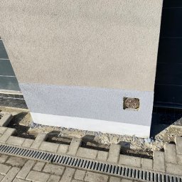 Usługi Budowlane Artur Ochodek - Fragment elewacji budynku z widocznym ociepleniem, cokół pomalowany na szaro, obok ułożona kostka brukowa i kratka odpływowa.