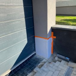 Usługi Budowlane Artur Ochodek - Przygotowanie do ocieplenia narożnika budynku z użyciem pomarańczowej taśmy maskującej i folii ochronnej, widoczna siatka zbrojeniowa przy garażu oraz kostka brukowa z odpływem liniowym.