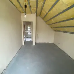 Poddasze z ocieplonym stropem wełną mineralną, widoczna konstrukcja nośna i prowizoryczne oświetlenie żarówką, w tle widoczne rusztowanie w przejściu do innego pomieszczenia.