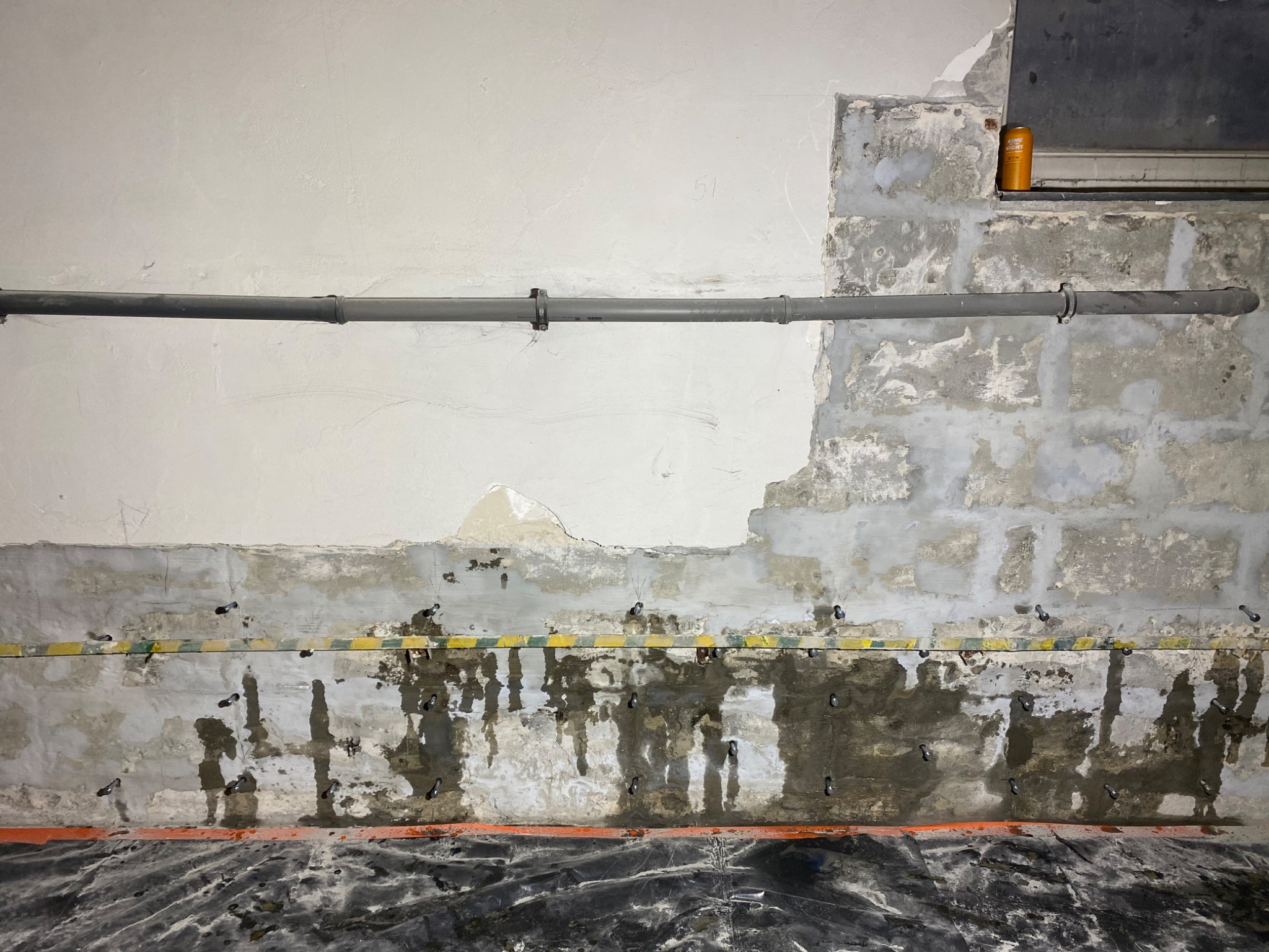 Ściana w trakcie przygotowania do prac wykończeniowych, widoczne uszkodzenia, rury instalacyjne, zabezpieczenia folią i żółto-czarna taśma. U góry puszka z napojem na tle okna.