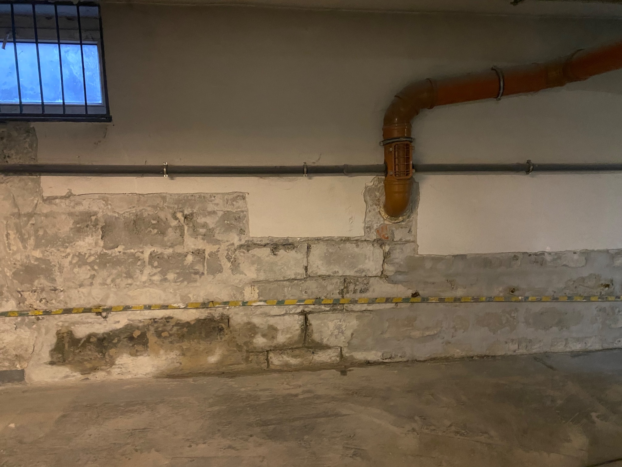 Fragment ściany z odsłoniętą cegłą i częściowo nałożonym tynkiem, widoczna rura kanalizacyjna oraz małe okno z kratą
