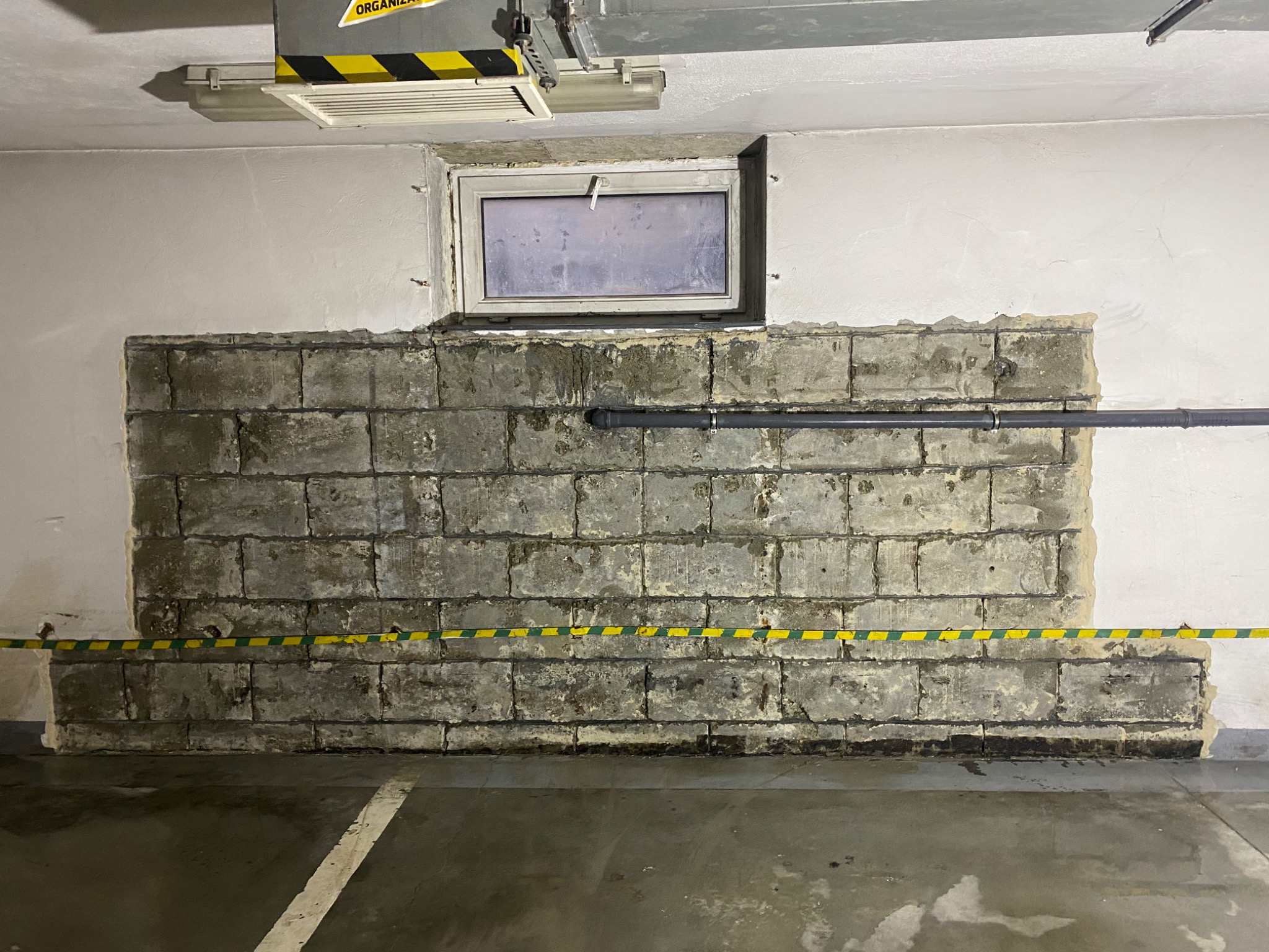 Fragment ściany z betonu komórkowego w garażu podziemnym, częściowo obłożony płytami, z widoczną instalacją rurową i taśmą ostrzegawczą.