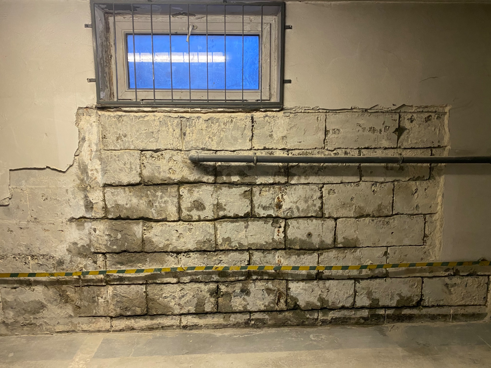 Odsłonięta, surowa ściana z bloków w pomieszczeniu w trakcie remontu, z widocznymi śladami wilgoci, rurą i taśmą ostrzegawczą, okno z kratą w tle.