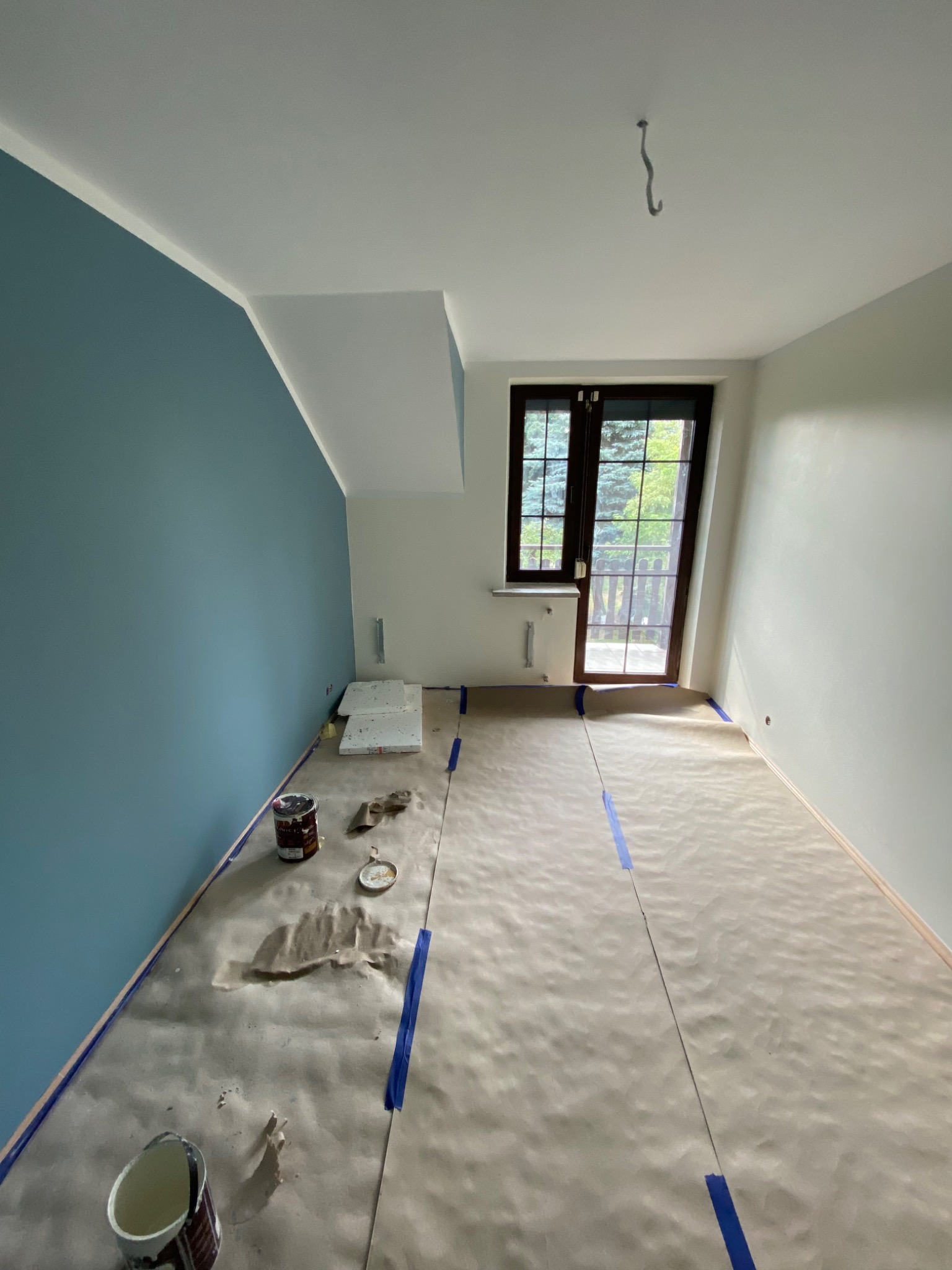 Pokój w trakcie malowania: jedna ściana pomalowana na niebiesko, podłoga zabezpieczona papierem malarskim, widoczne puszki z farbą i akcesoria malarskie. Na suficie widoczny hak.