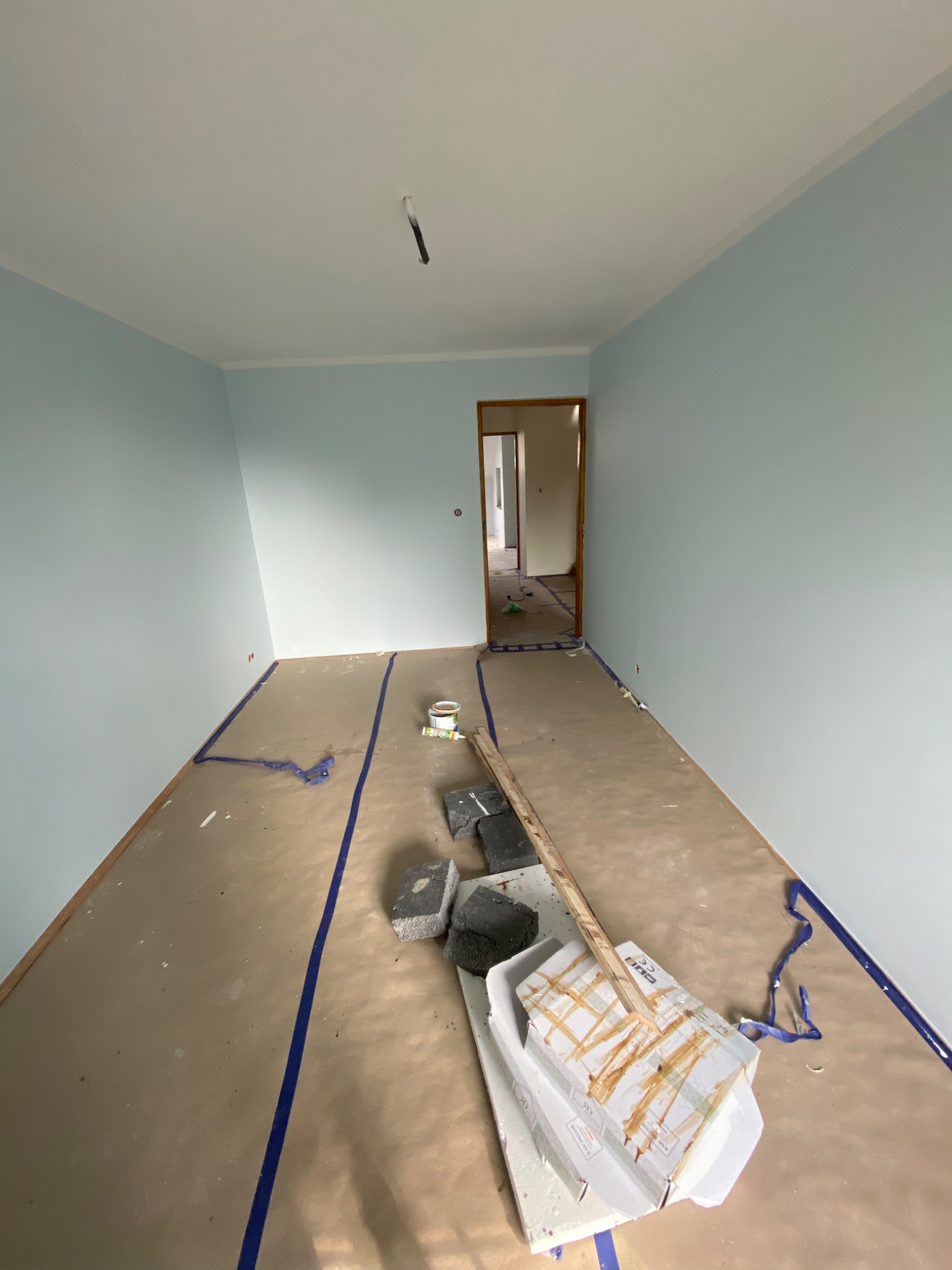 Pokój w trakcie malowania na jasny, błękitny odcień, podłoga zabezpieczona papierem malarskim, widoczne narzędzia i materiały malarskie, w tle otwarte drzwi do kolejnego pomieszczenia.