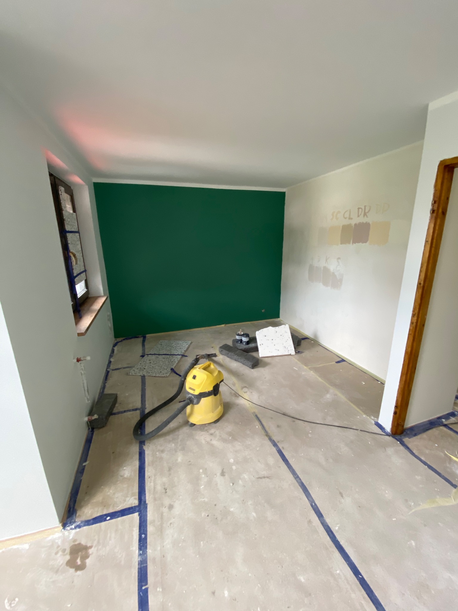 Pokój w trakcie prac: zielona ściana, próbki farb na sąsiedniej ścianie, zabezpieczona podłoga taśmą, odkurzacz przemysłowy i fragmenty płytek podłogowych.