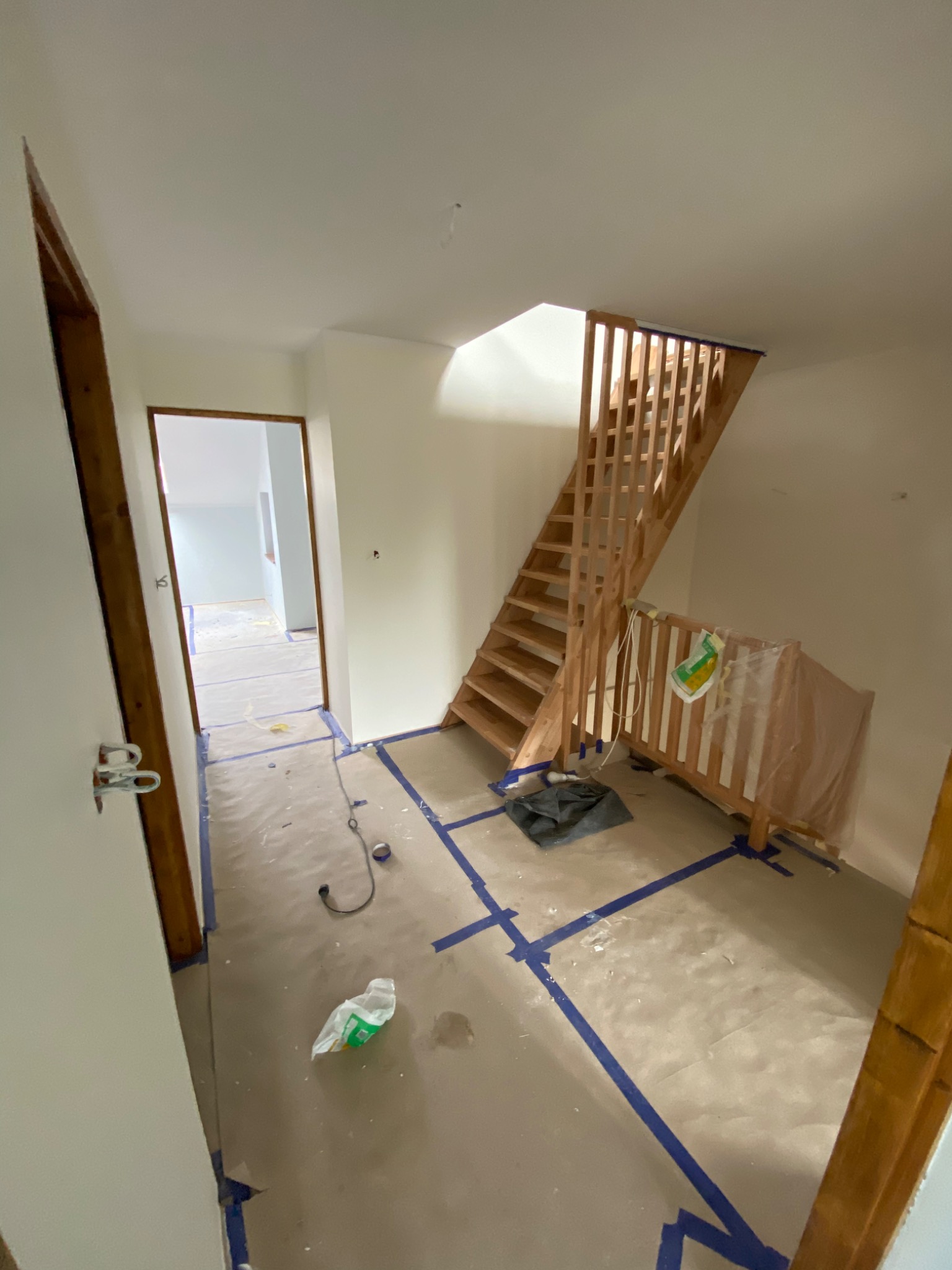 Wnętrze w trakcie malowania: drewniane schody, zabezpieczona podłoga z niebieską taśmą malarską, widoczne narzędzia i materiały malarskie.