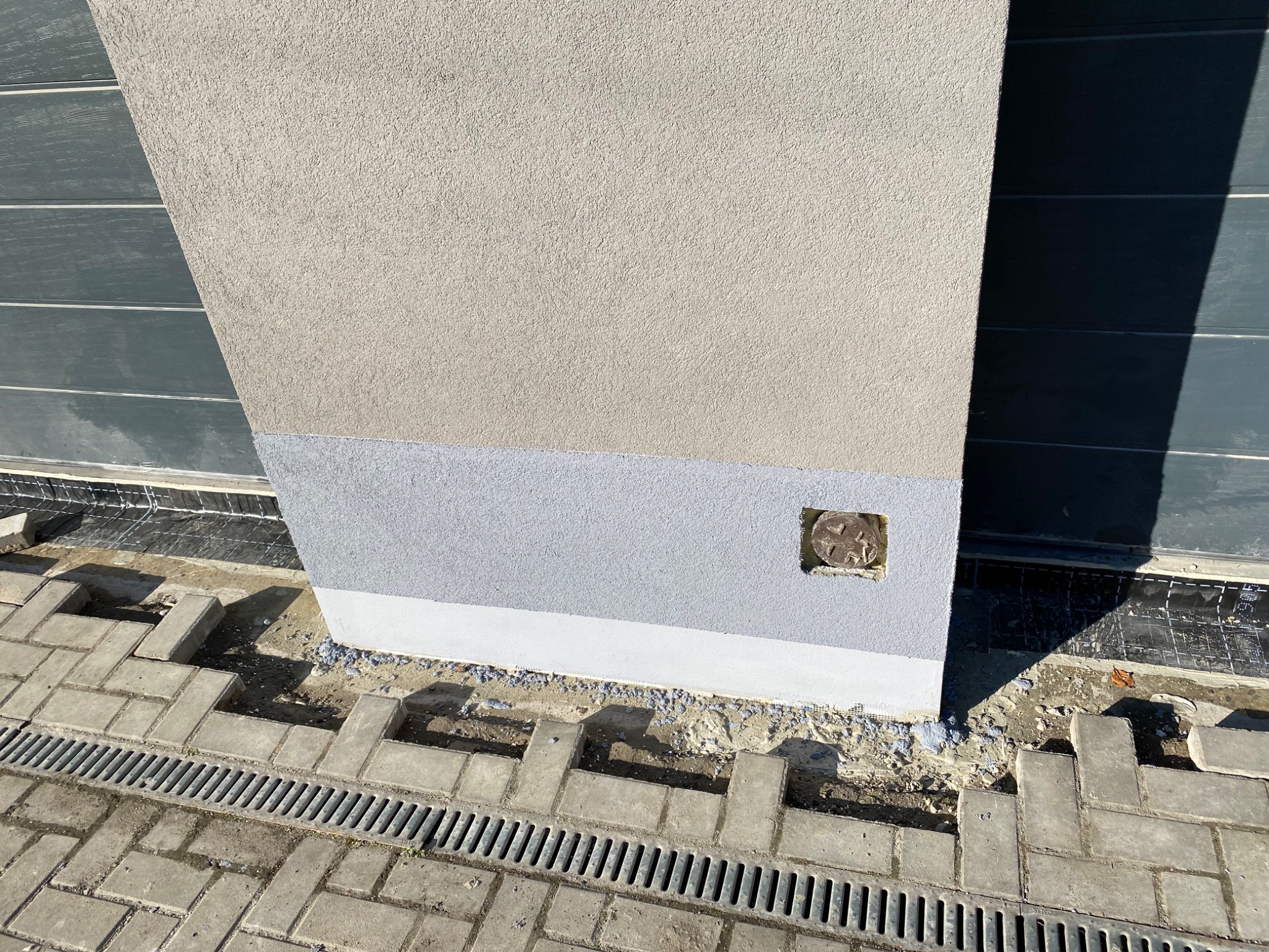 Fragment elewacji budynku z widocznym ociepleniem, cokół pomalowany na szaro, obok ułożona kostka brukowa i kratka odpływowa.