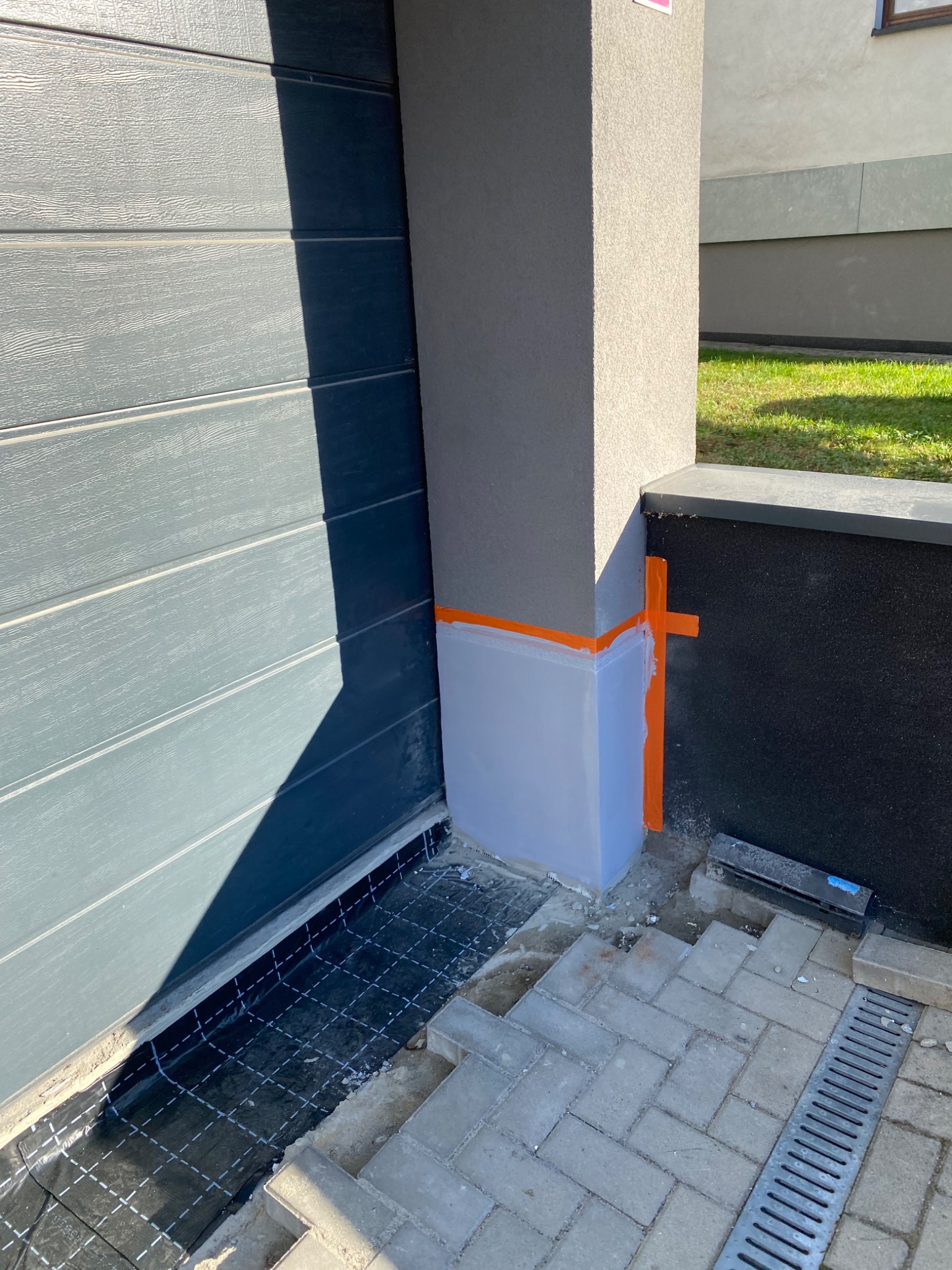 Przygotowanie do ocieplenia narożnika budynku z użyciem pomarańczowej taśmy maskującej i folii ochronnej, widoczna siatka zbrojeniowa przy garażu oraz kostka brukowa z odpływem liniowym.