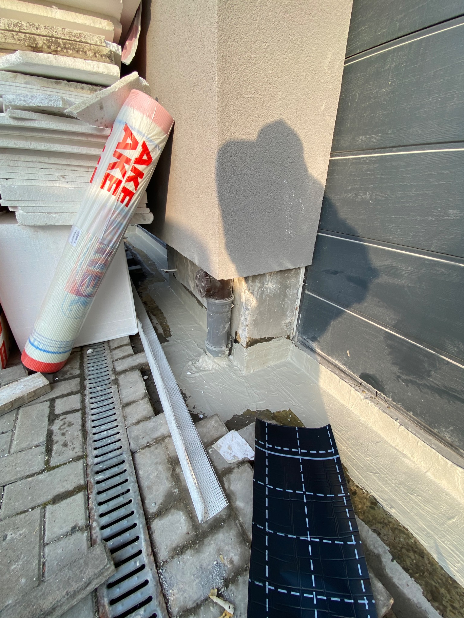 Ocieplanie fundamentów domu: rolka wełny mineralnej, siatka elewacyjna i fragment papy bitumicznej przy podstawie otynkowanej ściany budynku z szarymi panelami.