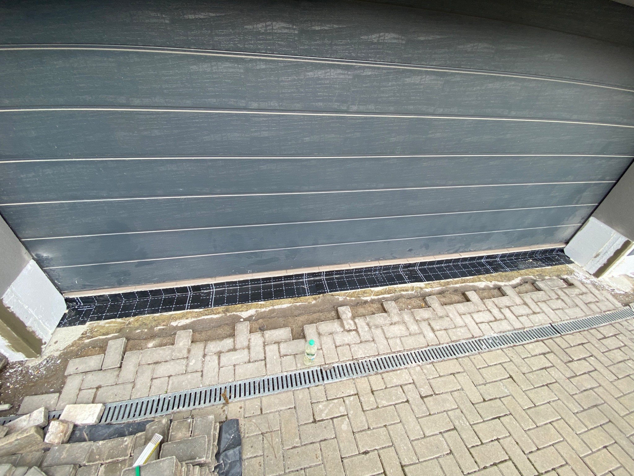 Montaż kostki brukowej przy garażu z odpływem liniowym, widoczne niedokończone fragmenty i zapas kostki.