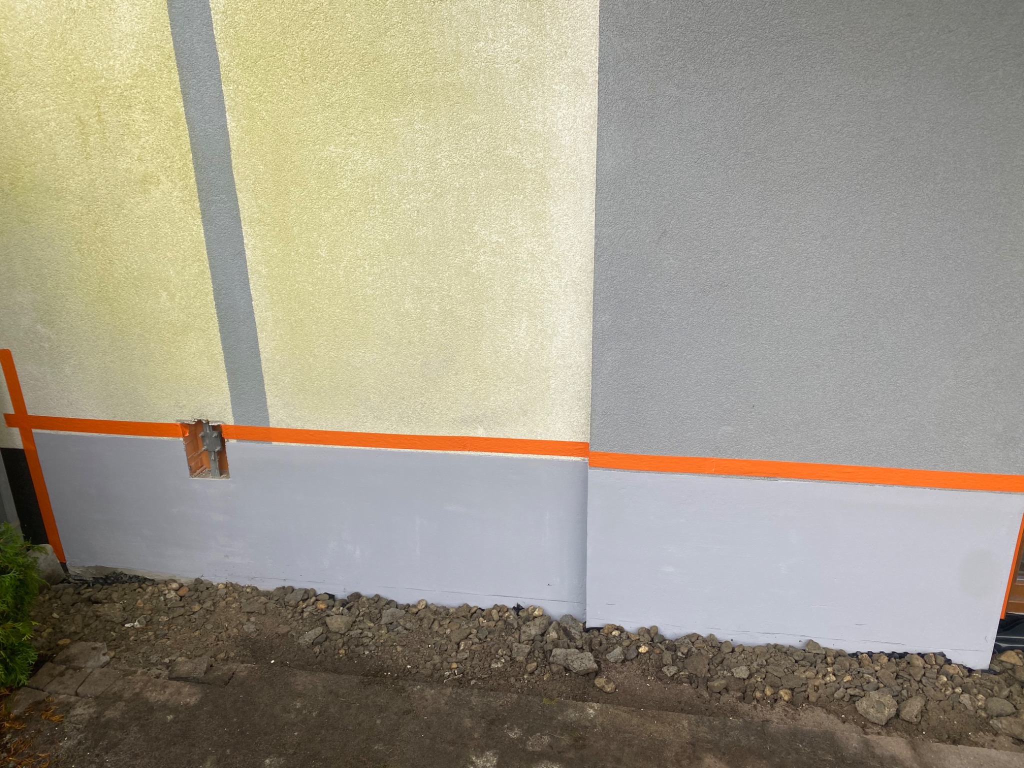 Fragment ściany budynku z widocznym ociepleniem w trakcie prac, z zastosowanym tynkiem w kolorze beżowym i szarym, oddzielonym pomarańczową taśmą malarską.