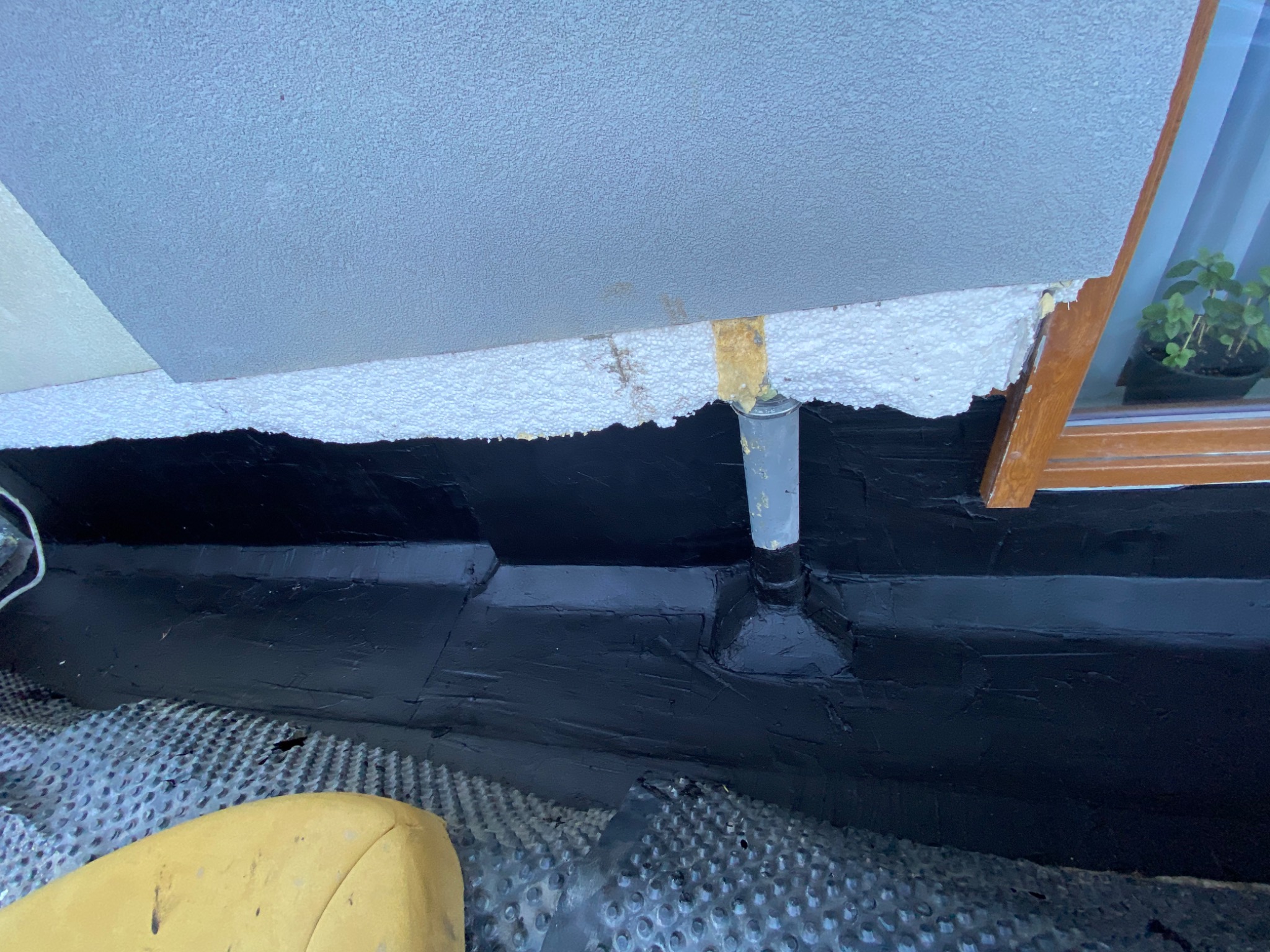 Izolacja przeciwwodna fundamentów z widoczną folią kubełkową, rurą odpływową i pianką montażową przy oknie piwnicznym.