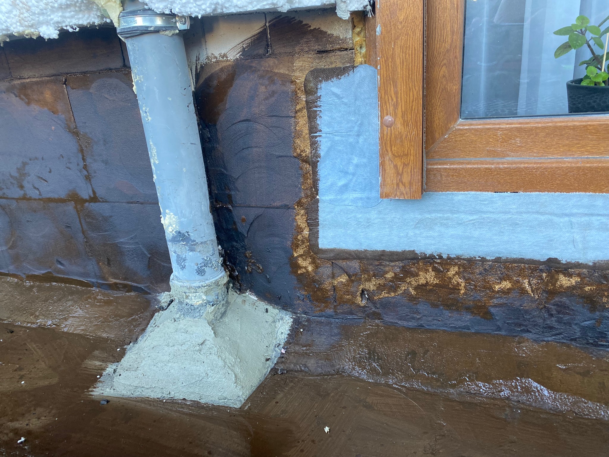 Ocieplanie ściany zewnętrznej przy oknie i rurze spustowej, widoczne fragmenty styropianu i świeżo nałożona warstwa izolacyjna.