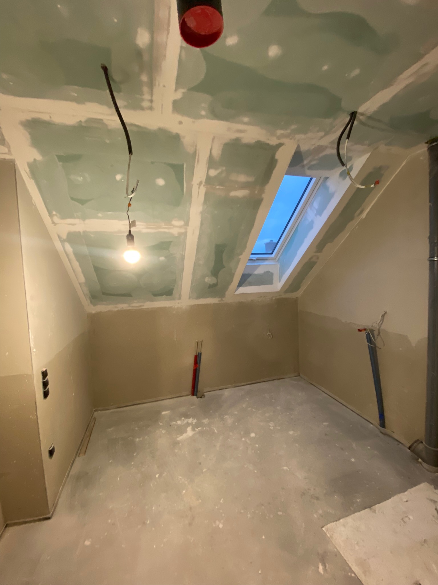 Poddasze w trakcie remontu: surowy stan z oknem dachowym, widoczne płyty kartonowo-gipsowe na suficie, instalacja elektryczna i rury wentylacyjne.