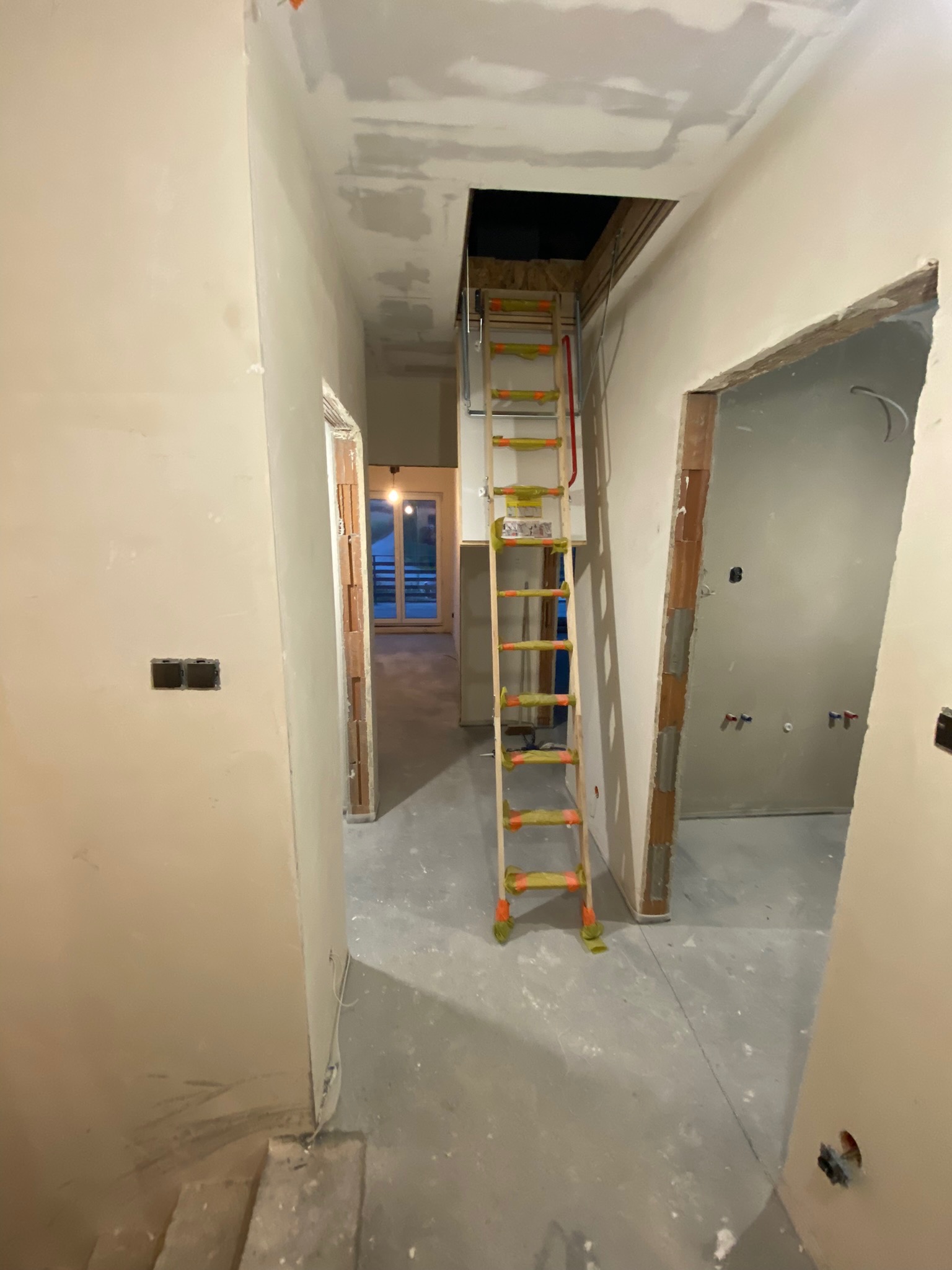 Wnętrze w trakcie prac przygotowawczych do wykończenia: ściany po gruntowaniu, widoczne otwory na gniazdka elektryczne, rozkładana drabina aluminiowa prowadząca do otworu w suficie na poddasze.