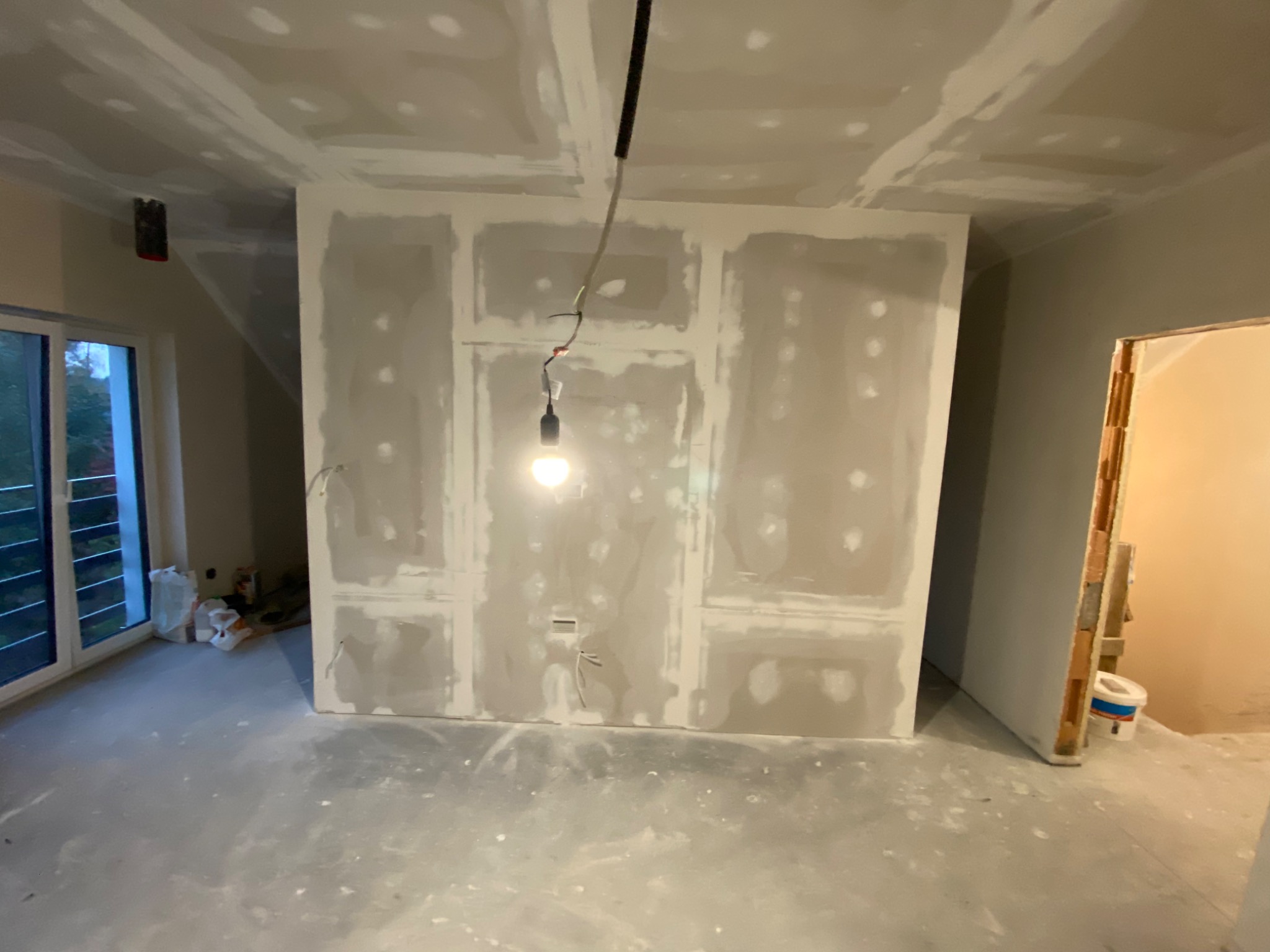 Surowy pokój z oknem i ścianami z płyt gipsowo-kartonowych w trakcie szpachlowania, wisząca żarówka oświetla wnętrze.