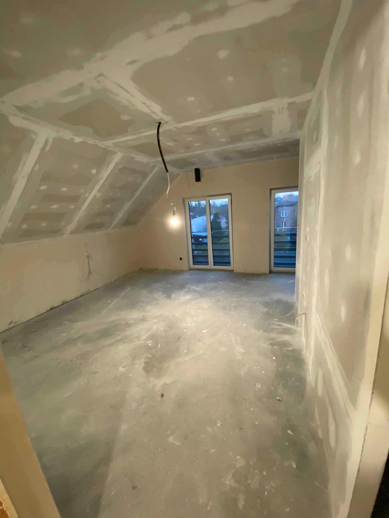 Poddasze po szpachlowaniu, widoczne ślady po gładzi na ścianach i suficie, przygotowane do dalszych prac wykończeniowych, z oknami balkonowymi wychodzącymi na zewnątrz.