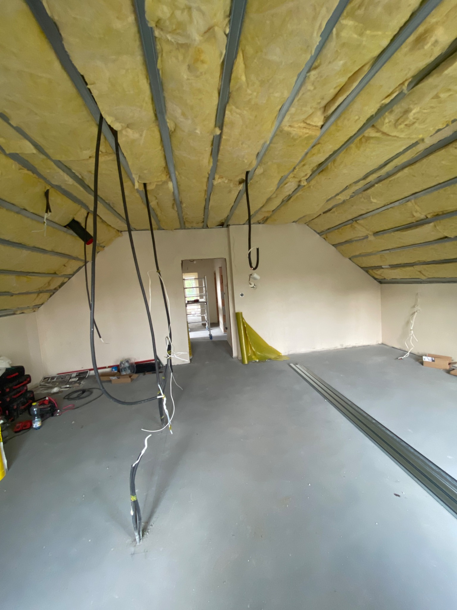 Poddasze w trakcie izolacji: widoczna wełna mineralna między metalowymi profilami sufitu, zwisające przewody elektryczne, surowy beton na podłodze, narzędzia w tle.