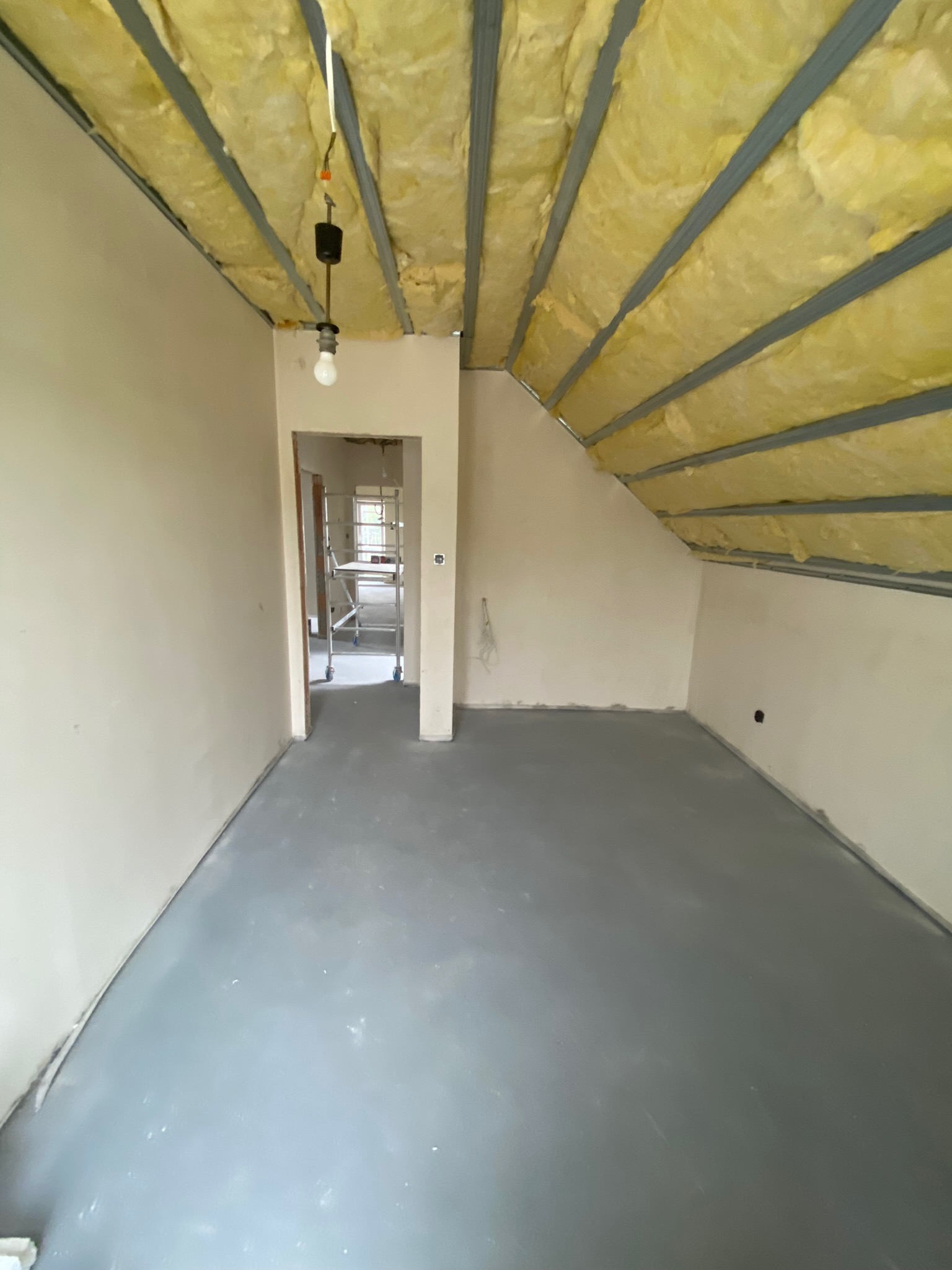 Poddasze z ocieplonym stropem wełną mineralną, widoczna konstrukcja nośna i prowizoryczne oświetlenie żarówką, w tle widoczne rusztowanie w przejściu do innego pomieszczenia.