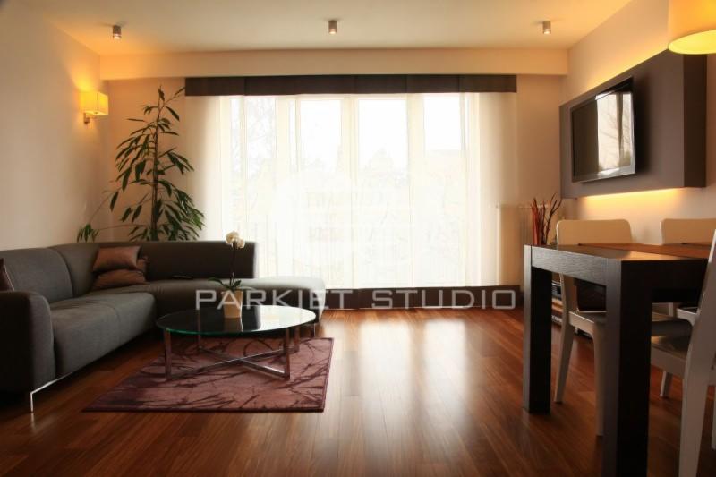 Salon z ciemną, błyszczącą podłogą z egzotycznego drewna Afromozja, szary narożnik, szklany stolik kawowy na tle okna z jasnymi zasłonami.