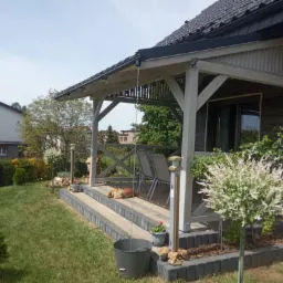 Zadaszony taras z drewnianą konstrukcją, schodami z kostki, śpiącym psem i ozdobnymi roślinami w ogrodzie.