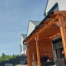 Zadaszona drewniana konstrukcja przylegająca do nowoczesnego domu z szarymi oknami, widok z niskiej perspektywy na tle błękitnego nieba.