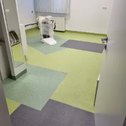 Wnętrze pomieszczenia z nową, wielokolorową podłogą ułożoną z kwadratowych płytek w odcieniach zieleni i szarości, z widocznym sprzętem owiniętym folią ochronną w tle.