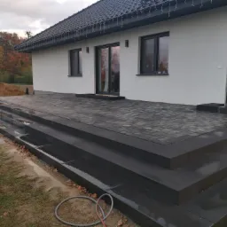 Nowoczesny taras z kostki brukowej w odcieniach szarości, prowadzący do domu z białymi ścianami i ciemnymi oknami, widoczny stos piasku obok