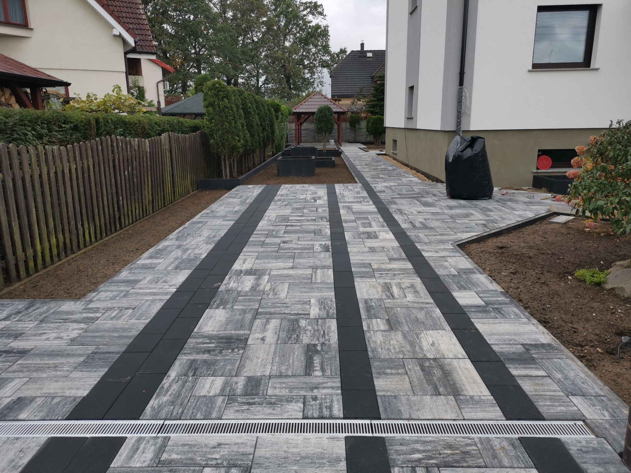 Ułożona ścieżka z kostki brukowej w odcieniach szarości, z czarnymi pasami i odpływem liniowym, prowadząca do altany w ogrodzie, widok z perspektywy pieszego.
