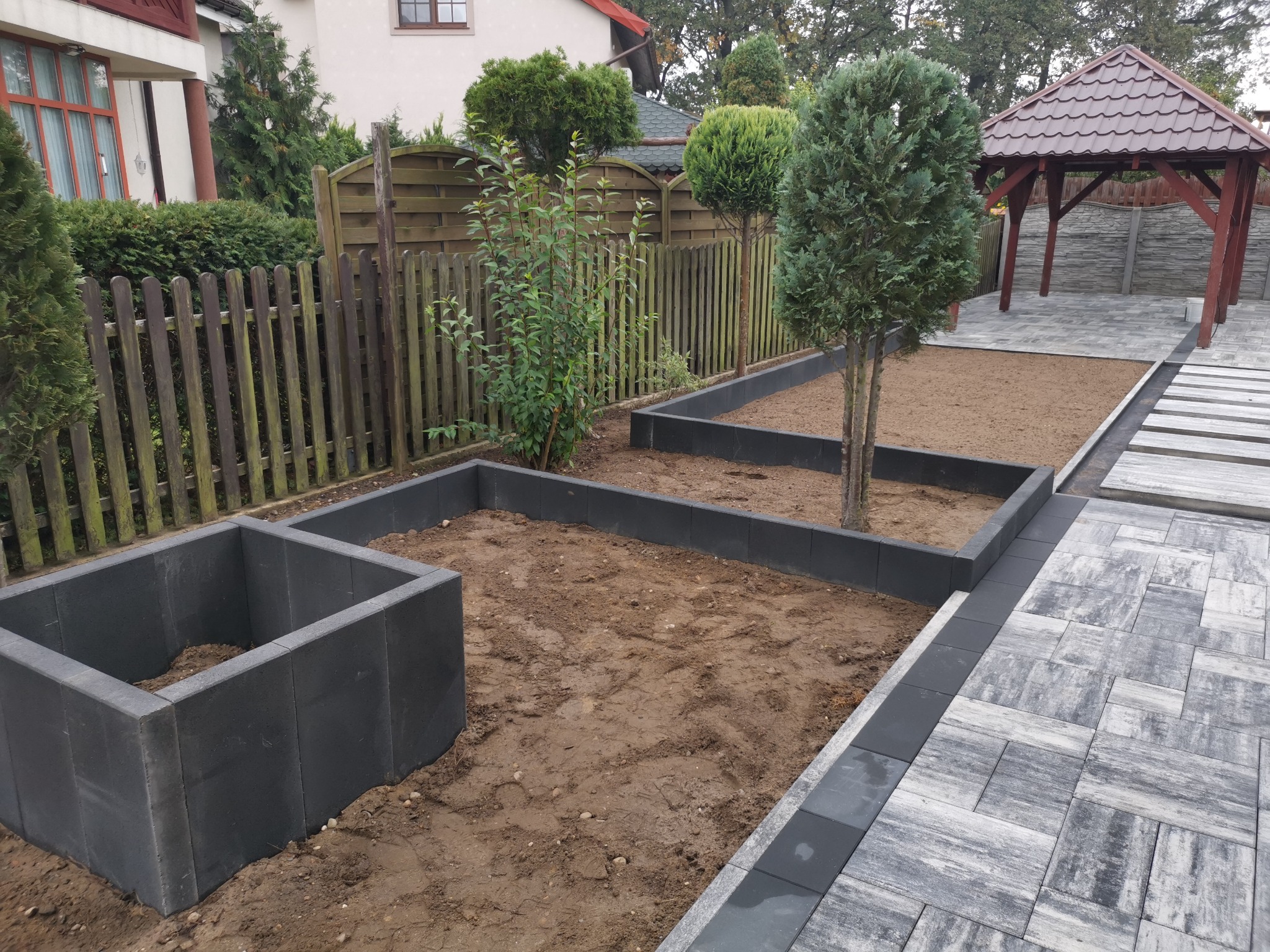 Przygotowanie podłoża pod rabaty kwiatowe z obrzeżami z szarej kostki betonowej, widoczny fragment ułożonej nawierzchni z kostki brukowej w odcieniach szarości oraz drewniana altana w ogrodzie.