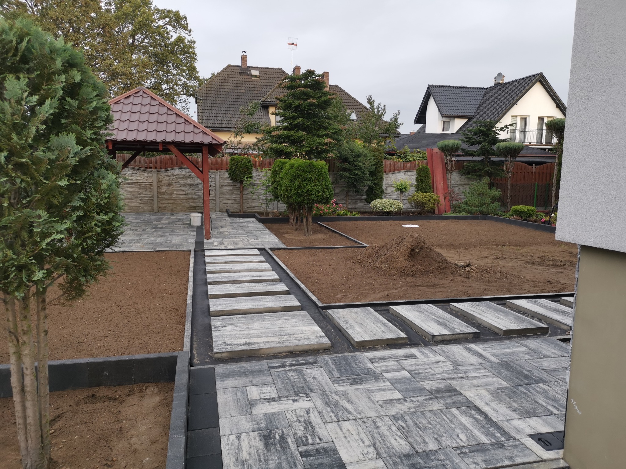 Nowo wyłożony, wzorzysty chodnik z szarych płyt betonowych i kostki, otaczający rabaty z ziemią i niskimi drzewkami, w tle altana i domy.