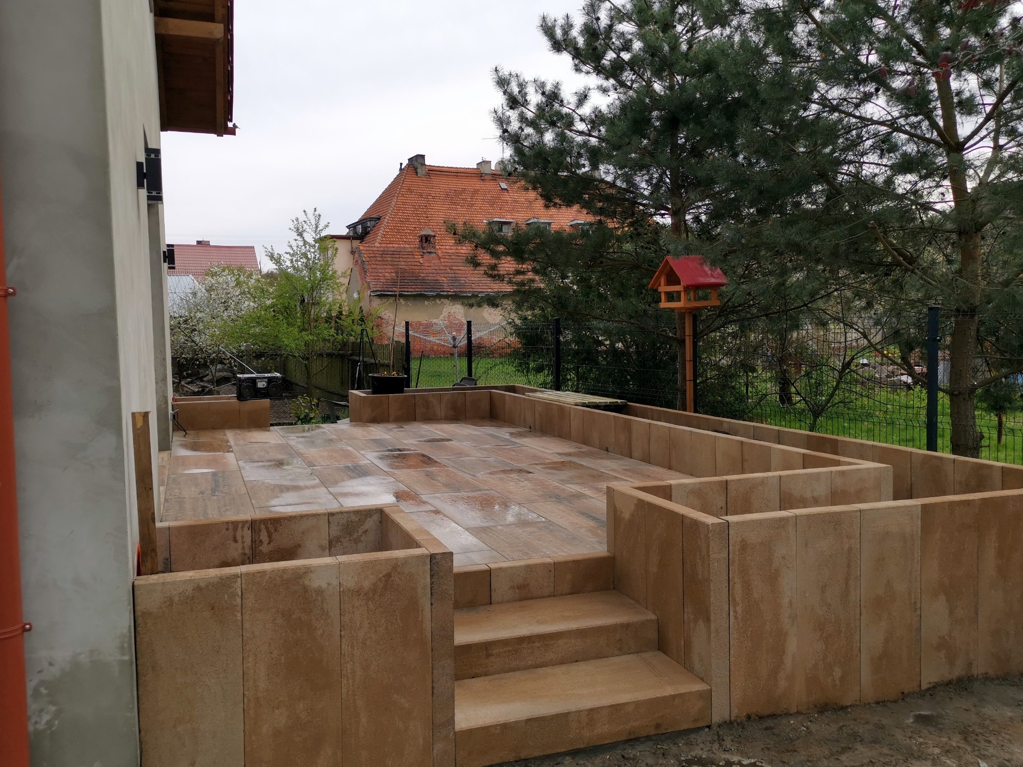 Ułożony taras z kostki brukowej z niskimi ściankami i schodami, widok z boku, w tle dom z czerwoną dachówką i karmnik dla ptaków.