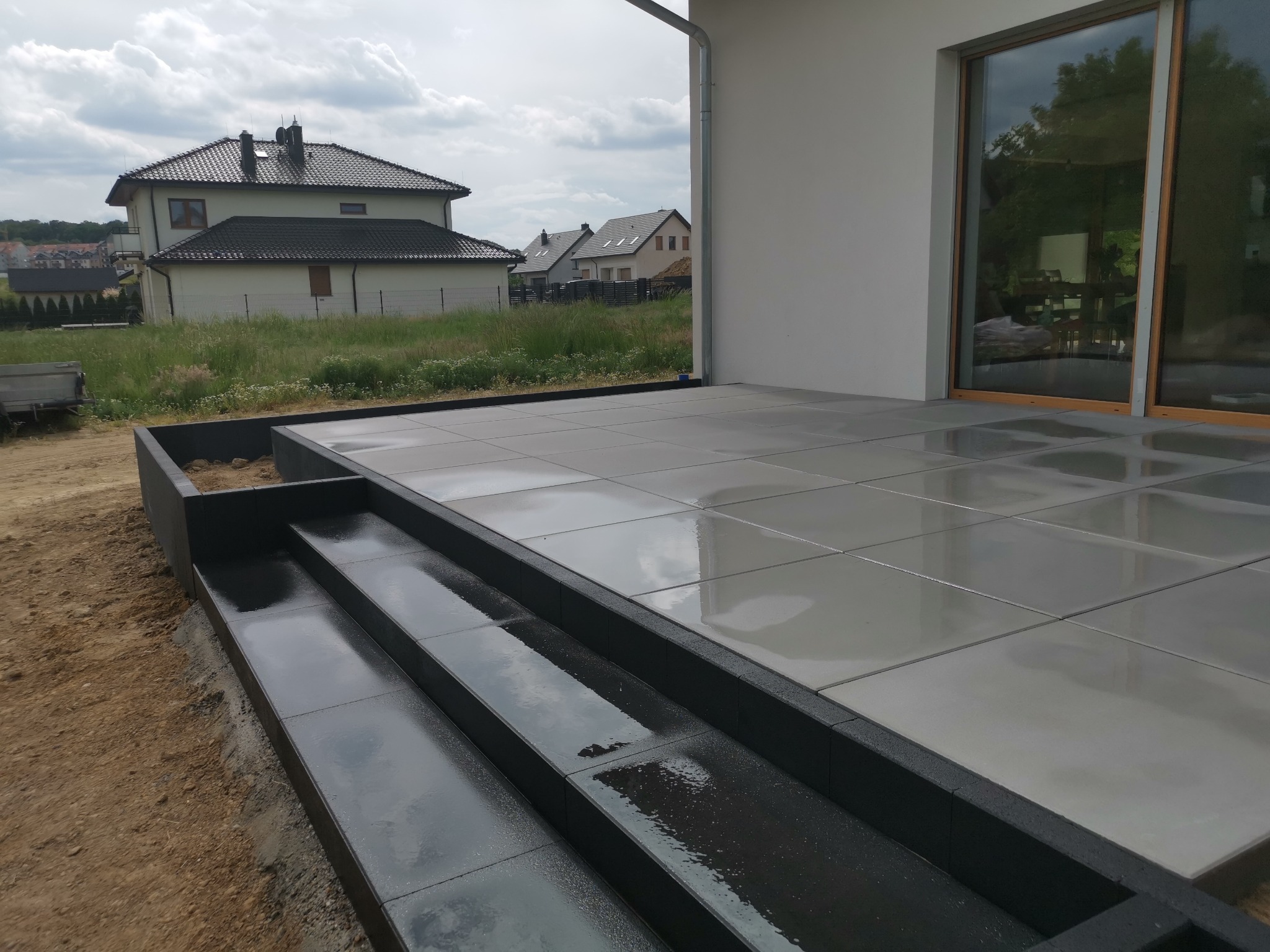 Nowoczesny taras i schody wyłożone szarymi płytami, widok na dom i okoliczną zabudowę w tle, pochmurny dzień.