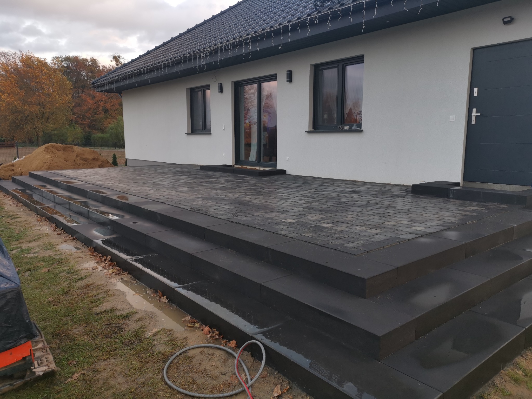 Nowoczesny taras z kostki brukowej w odcieniach szarości, prowadzący do domu z białymi ścianami i ciemnymi oknami, widoczny stos piasku obok