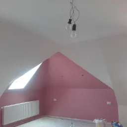 Pomalowane na różowo poddasze ze skosami, z grzejnikiem pod oknem dachowym i wiszącymi żarówkami na przewodach, w trakcie prac wykończeniowych.