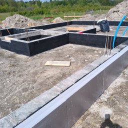 S-BUD SŁAWOMIR NIEMIEC - Zalane fundamenty domu z izolacją termiczną, widoczne wystające pręty zbrojeniowe i czarna izolacja przeciwwilgociowa. W tle ziemia i zieleń.