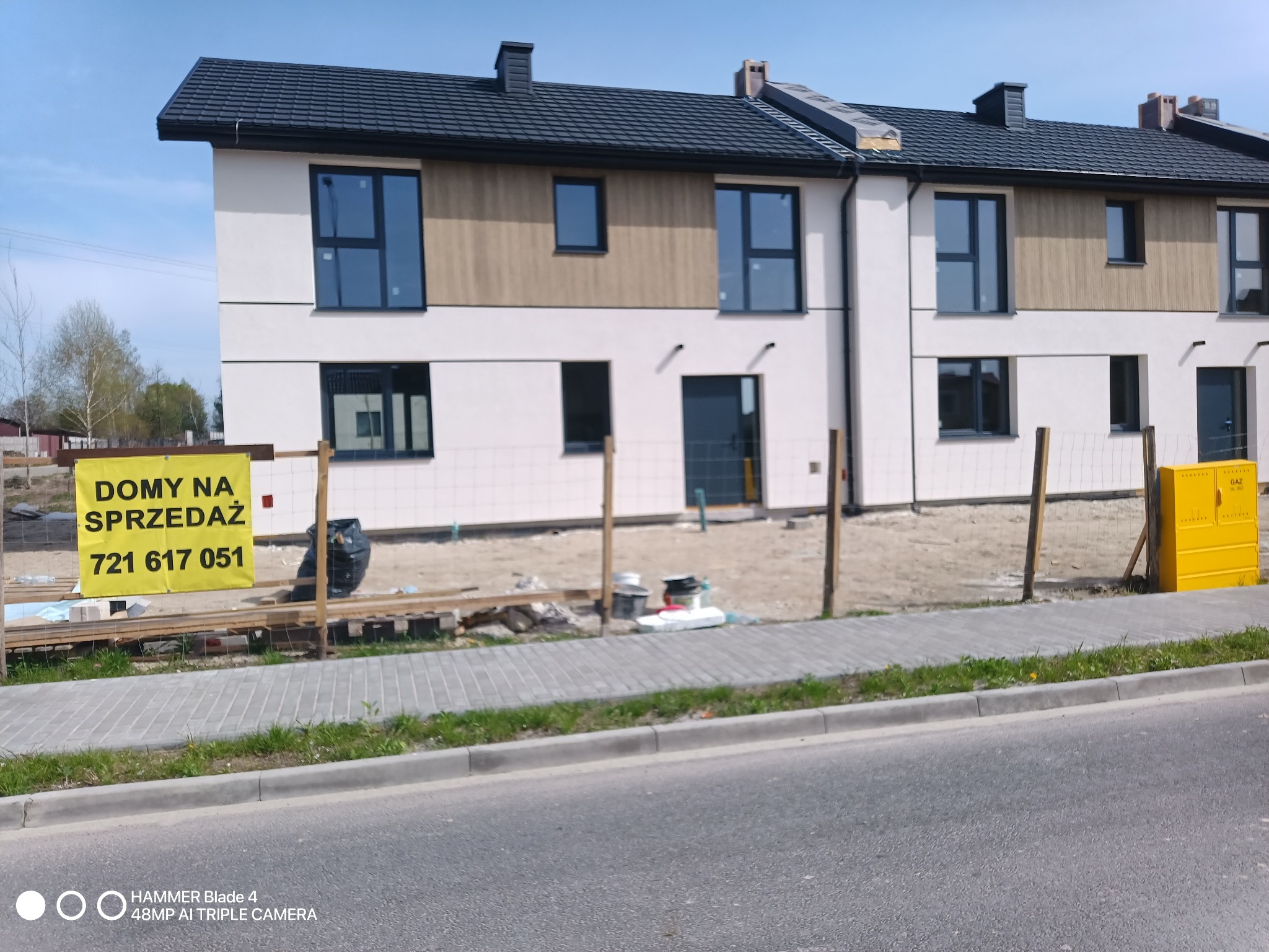 Nowoczesne domy szeregowe z białymi elewacjami i drewnianymi akcentami, widoczne z ulicy. Żółty baner 'Domy na sprzedaż' z numerem telefonu. Częściowo zagospodarowany teren.