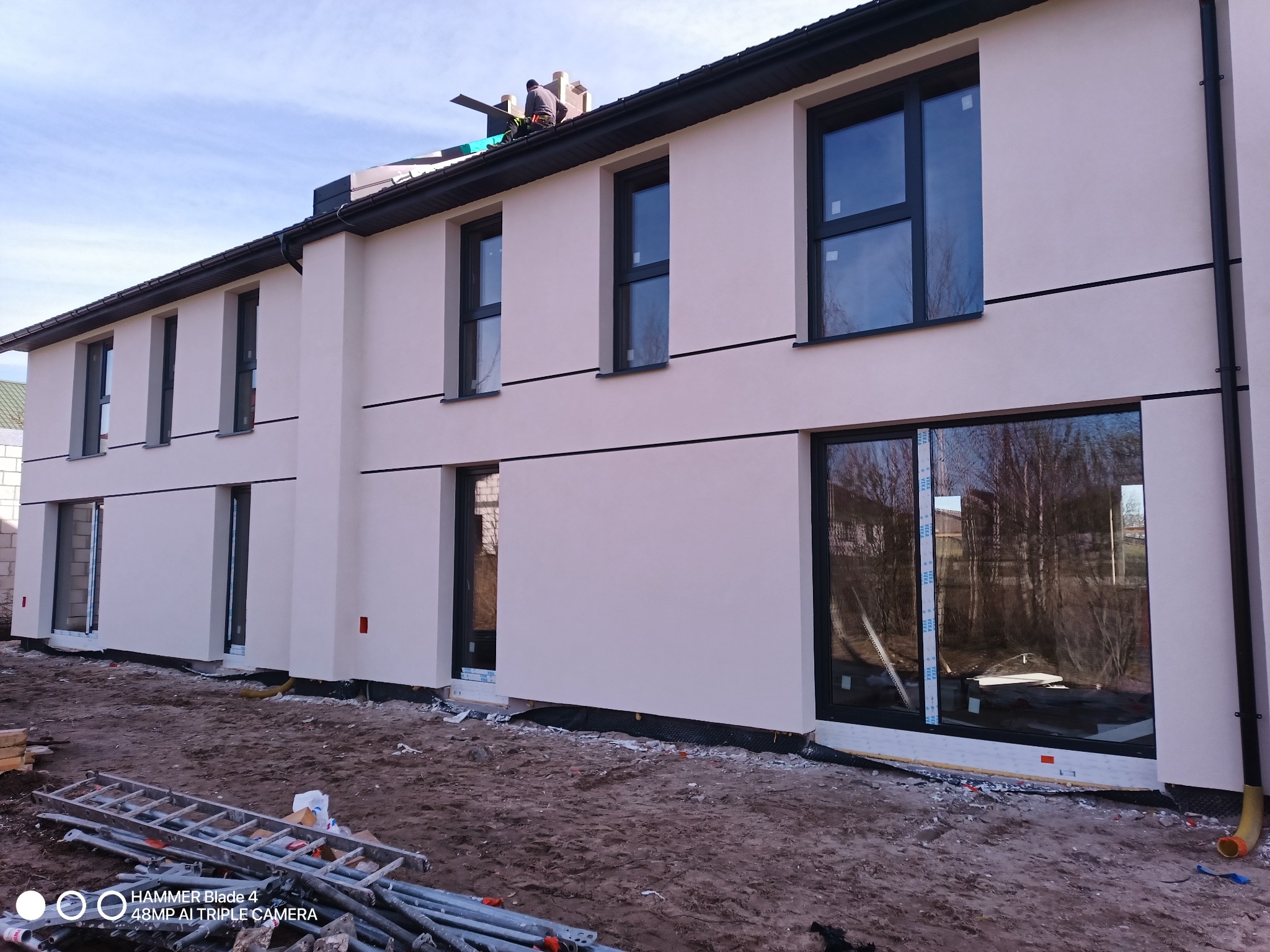 Nowoczesny dom z jasną elewacją i czarnymi oknami. Na dachu widoczny pracownik. Na pierwszym planie rusztowanie i drabina. Dom w trakcie wykończenia.