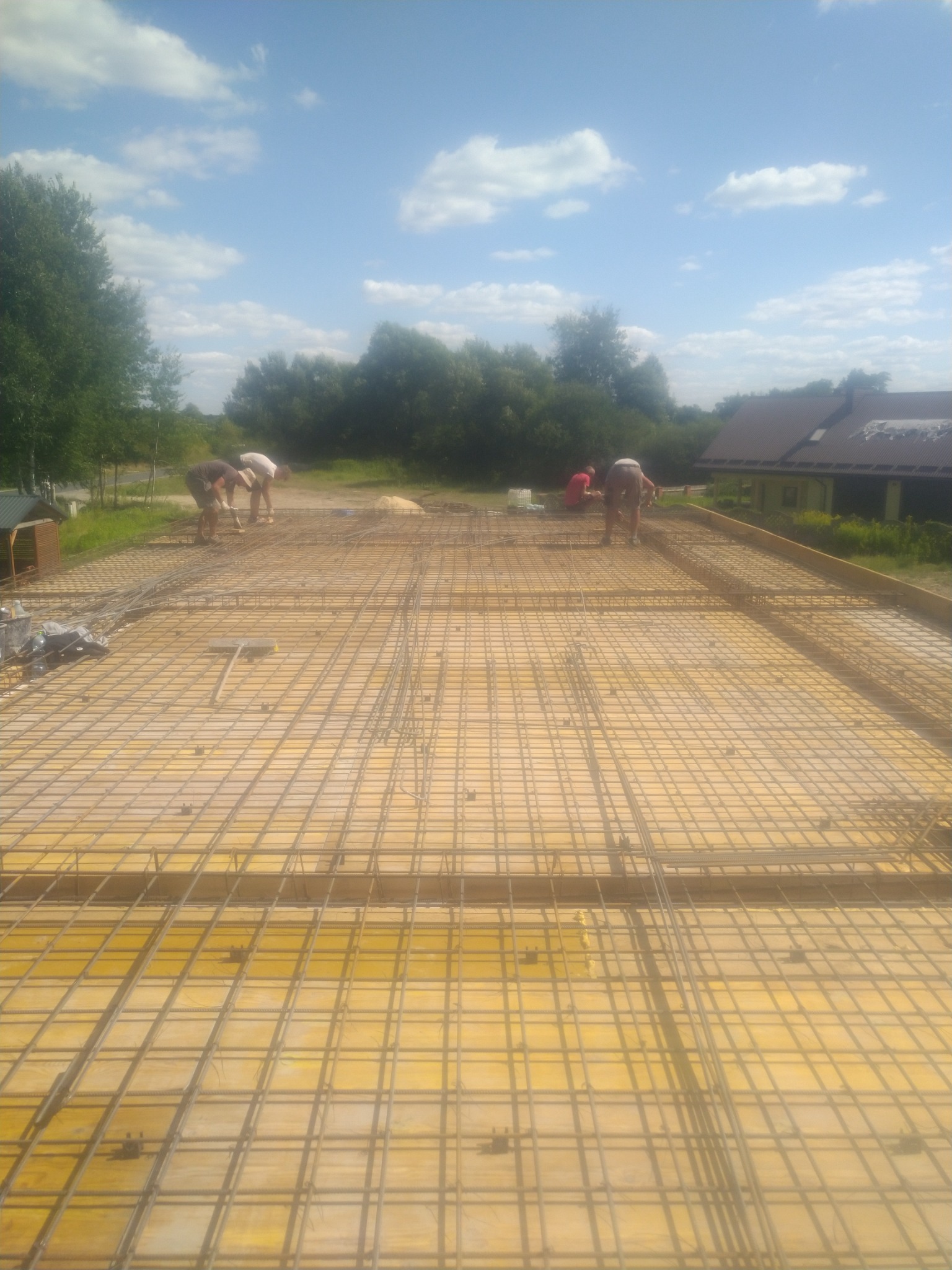 Zbrojenie fundamentów na żółtym szalunku, widoczni pracownicy w trakcie pracy, w tle zieleń i budynek. Ujęcie z góry, w słoneczny dzień.