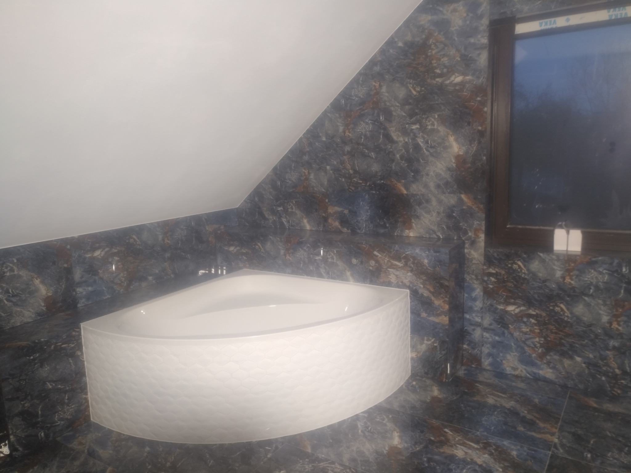 Wykończona łazienka na poddaszu z narożną wanną o fakturze plastra miodu i marmurowymi płytkami w odcieniach granatu i brązu, okno w ciemnej ramie.