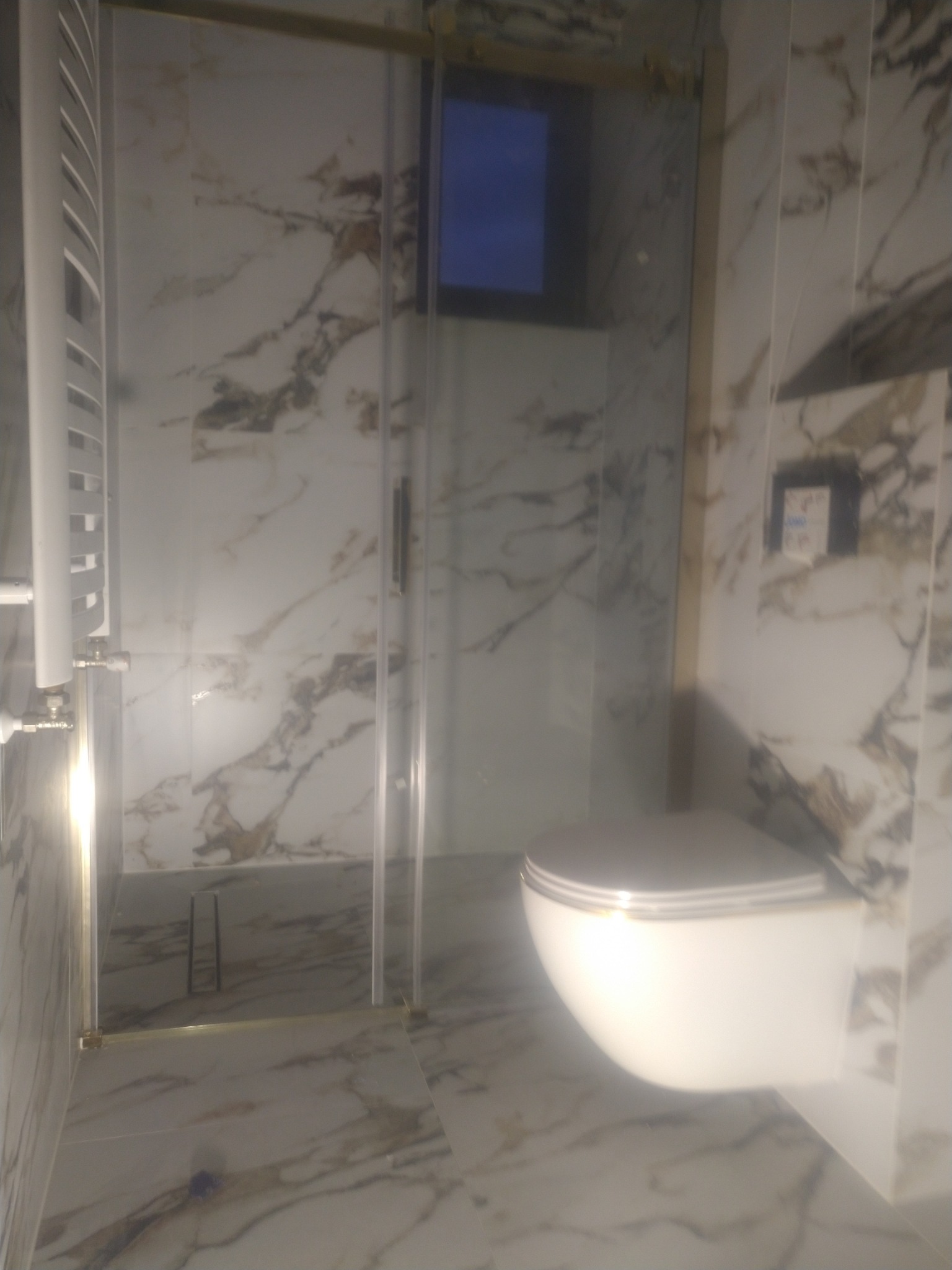 Wykończona łazienka z kabiną prysznicową i podwieszaną toaletą. Marmurowe płytki na ścianach i podłodze. Złote akcenty na kabinie i toalecie. Nowoczesny design.