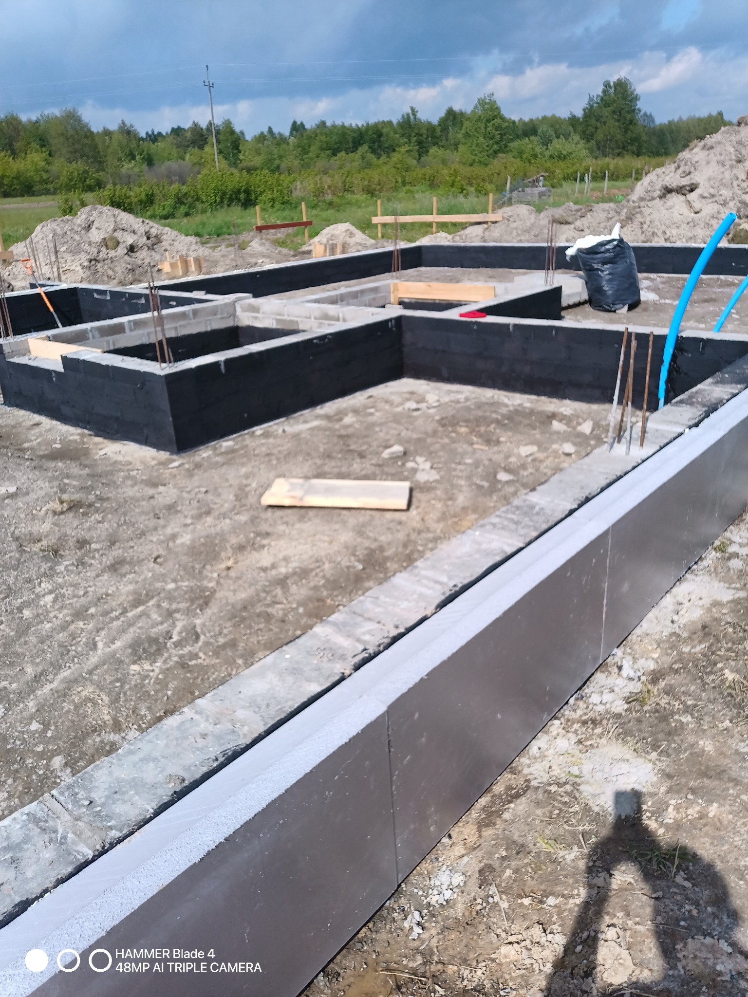 Zalane fundamenty domu z izolacją termiczną, widoczne wystające pręty zbrojeniowe i czarna izolacja przeciwwilgociowa. W tle ziemia i zieleń.