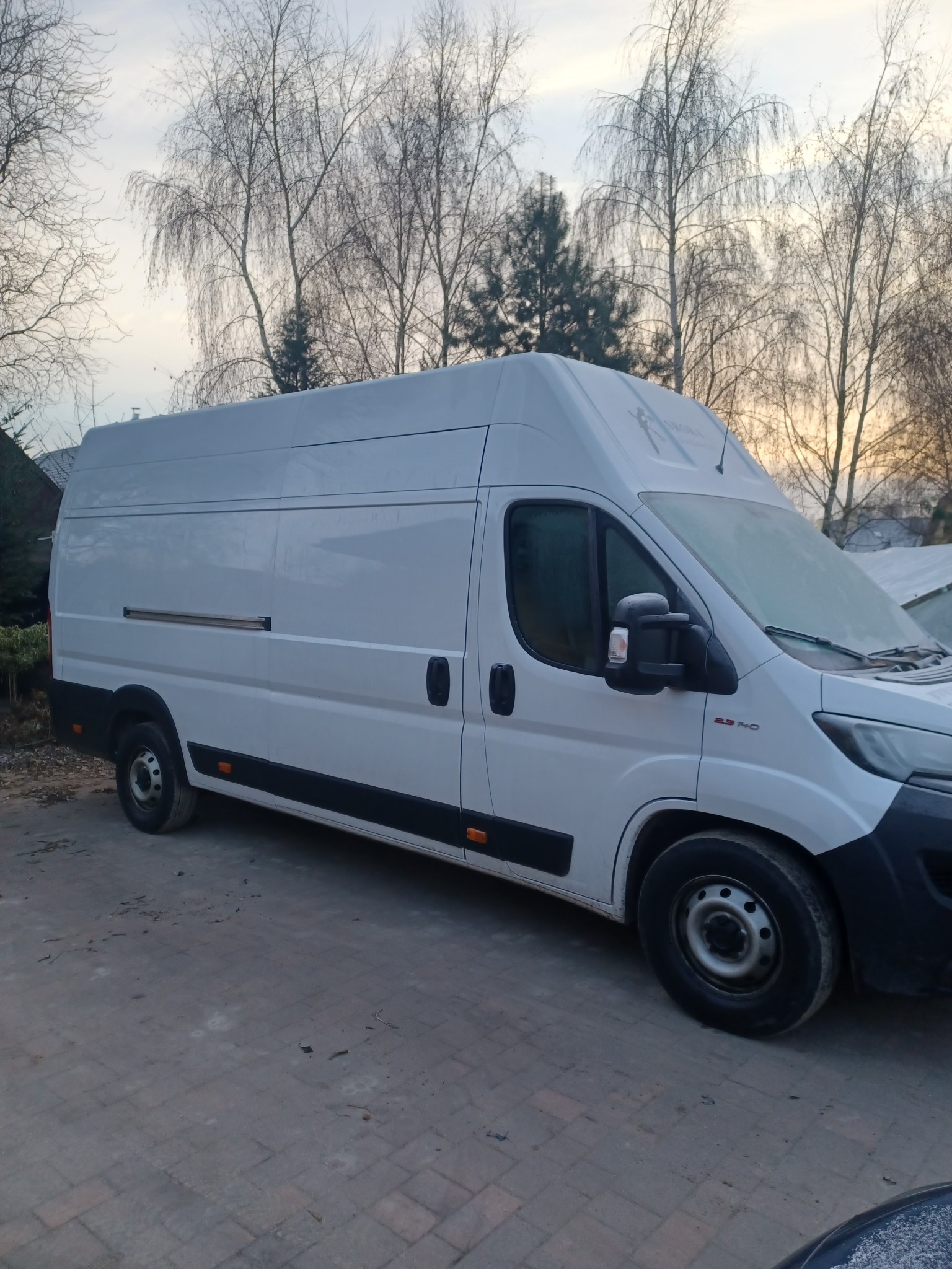 Biały bus dostawczy na brukowanym podjeździe w Poznaniu, z lekko oszronioną przednią szybą, na tle drzew bez liści i zachmurzonego nieba.